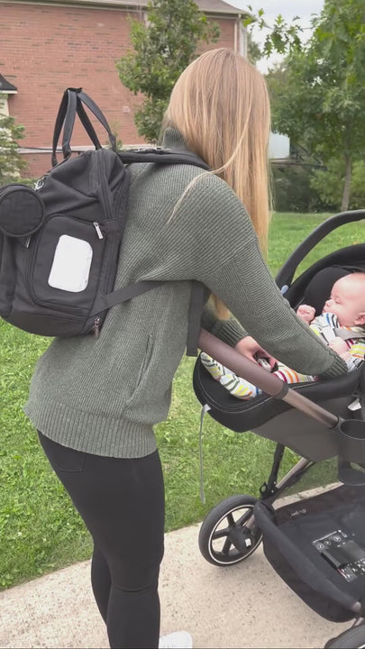 Video showing a women using a diaper bag with her baby. She opens the changing stations and places the baby on it for a nap while sitting on the grass. After she packs the diaper bag and attaches is to the stroller using the stroller straps.